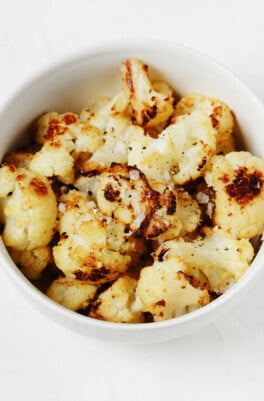 An overhead image of a white bowl, which is filled with roasted frozen cauliflower florets that are topped with flaky sea salt.