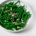 An image of a white ceramic bowl, which has been filled with brightly colored green beans and topped with sliced almonds and a vinaigrette.