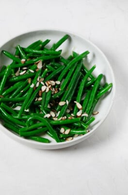 An image of a white ceramic bowl, which has been filled with brightly colored green beans and topped with sliced almonds and a vinaigrette.