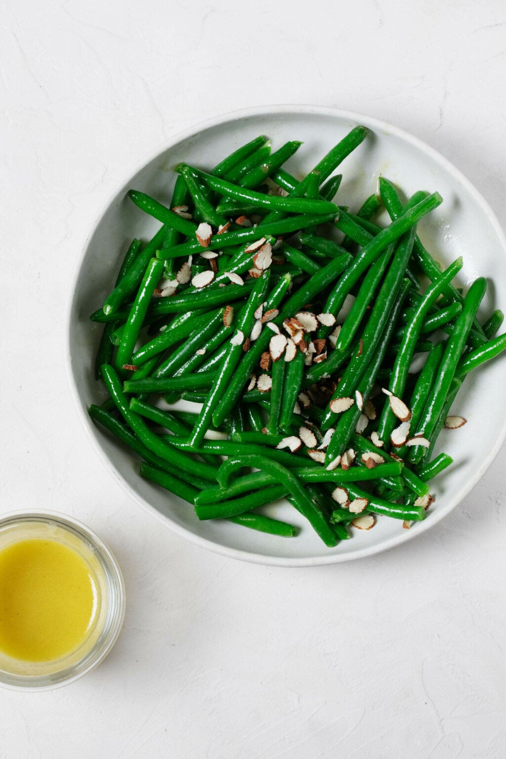An overhead image of steamed green beans in a white bowl, which are topped with a line of sliced almonds. A small bowl of dressing is situated nearby.