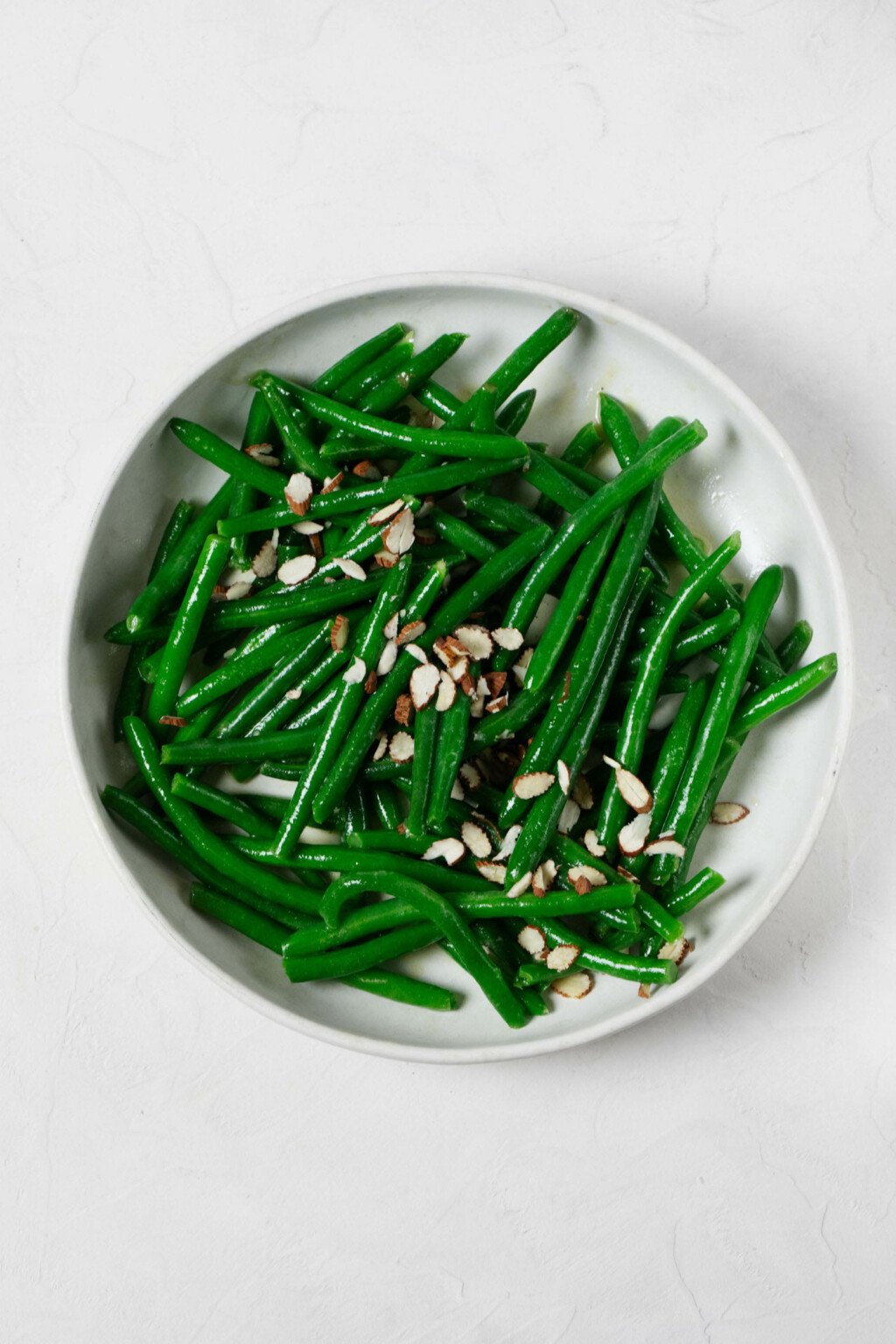 An overhead image of a bowl of steamed green beans, which are topped with a small row of sliced almonds.