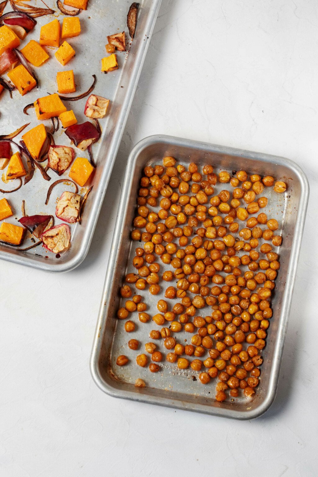 An overhead image of two silver baking sheets. One is used for roasting chickpeas, while the other has squash, onions, and apples.