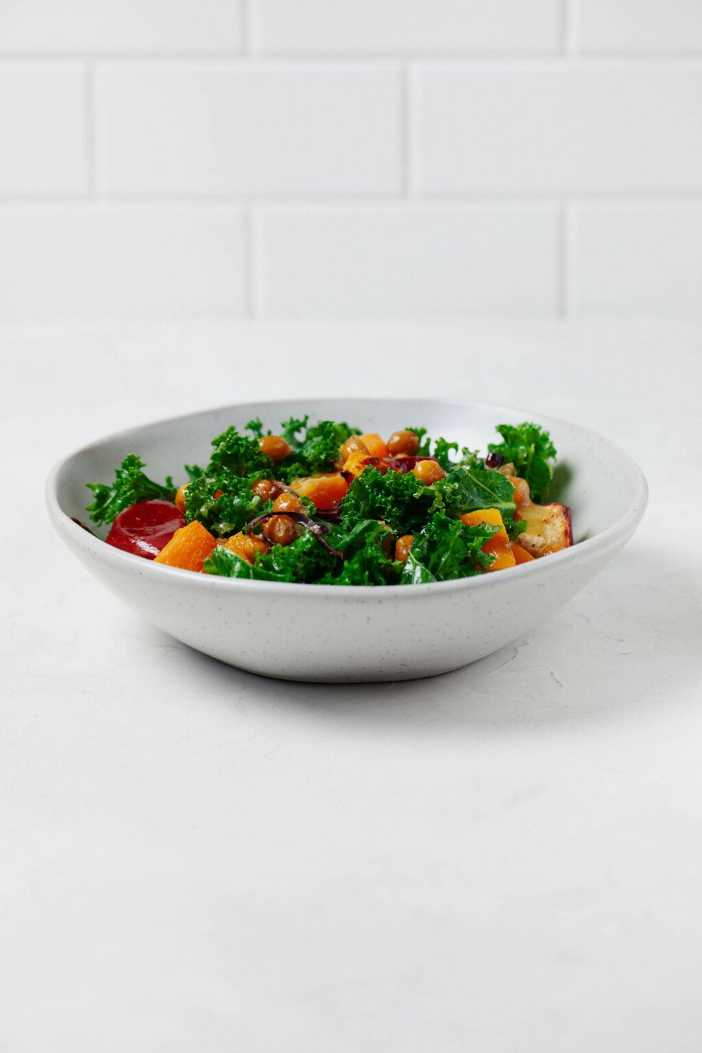 A shallow white bowl is pictured against a white tile backdrop. It contains a vibrant, green kale salad.