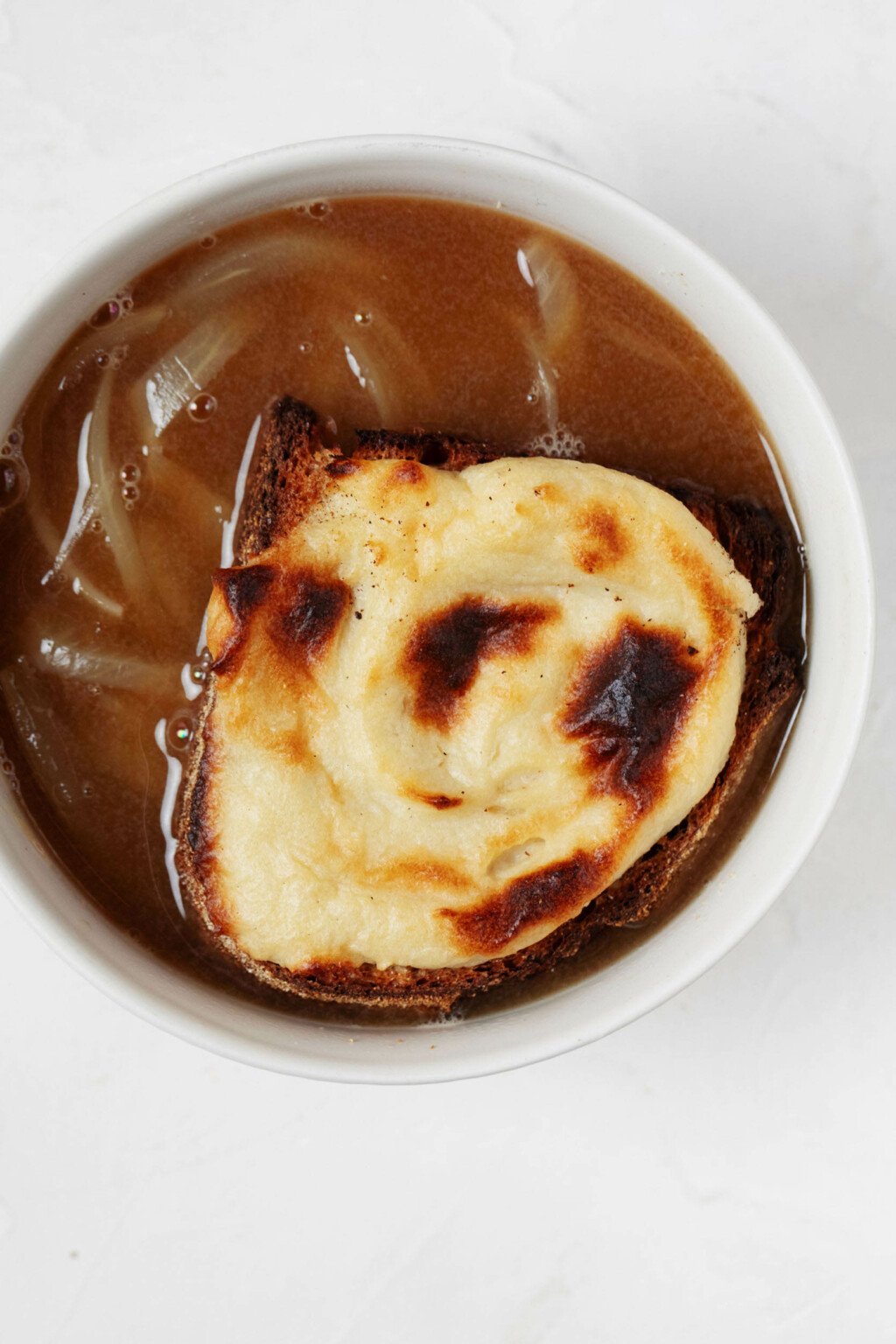 An overhead image of a round, white bowl of vegan French onion soup, which has been topped with a browned cheese toast. The toast rests directly in the bowl.