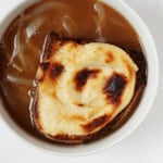 An overhead image of a round, white bowl of vegan French onion soup, which has been topped with a browned cheese toast. The toast rests directly in the bowl.