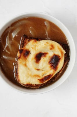 An overhead image of a round, white bowl of vegan French onion soup, which has been topped with a browned cheese toast. The toast rests directly in the bowl.