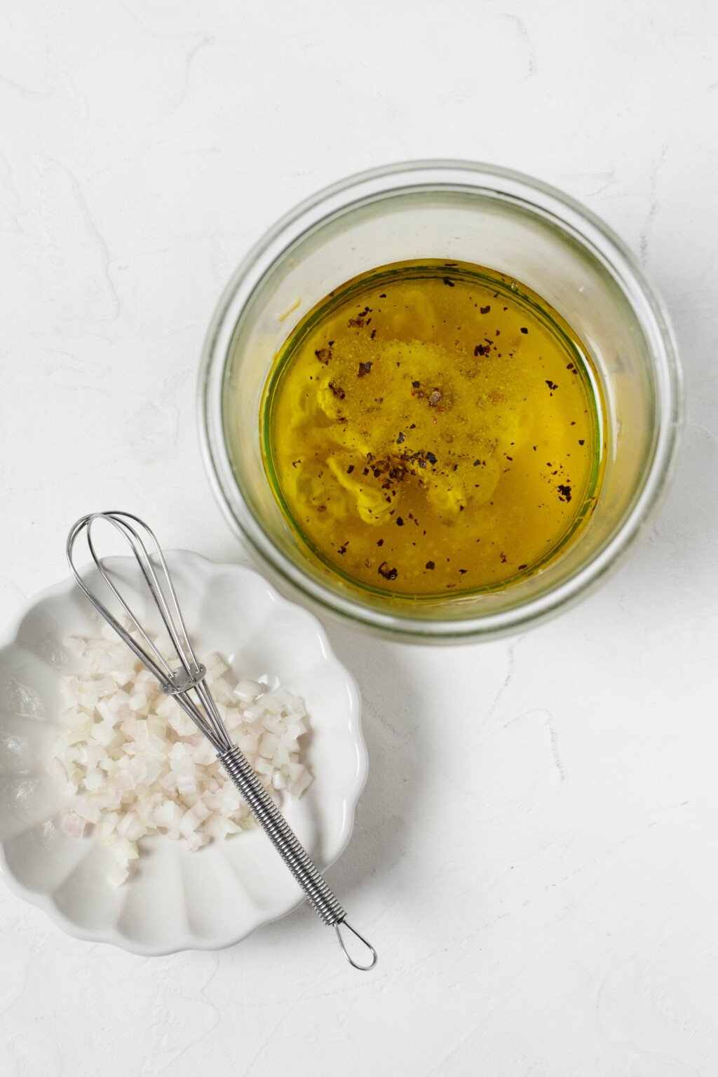 An overhead image of a small mason jar, which is resting next to a pinch bowl. The pinch bowl holds finely minced shallot and is pictured with a mini whisk.