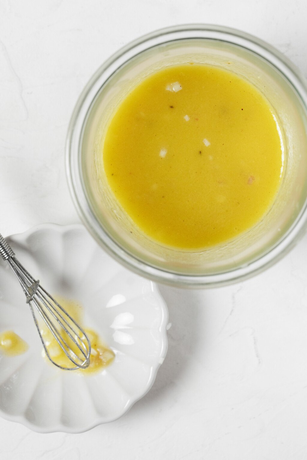 An overhead image of a pale yellow, vegan Champagne vinaigrette, with a tiny whisk resting nearby.