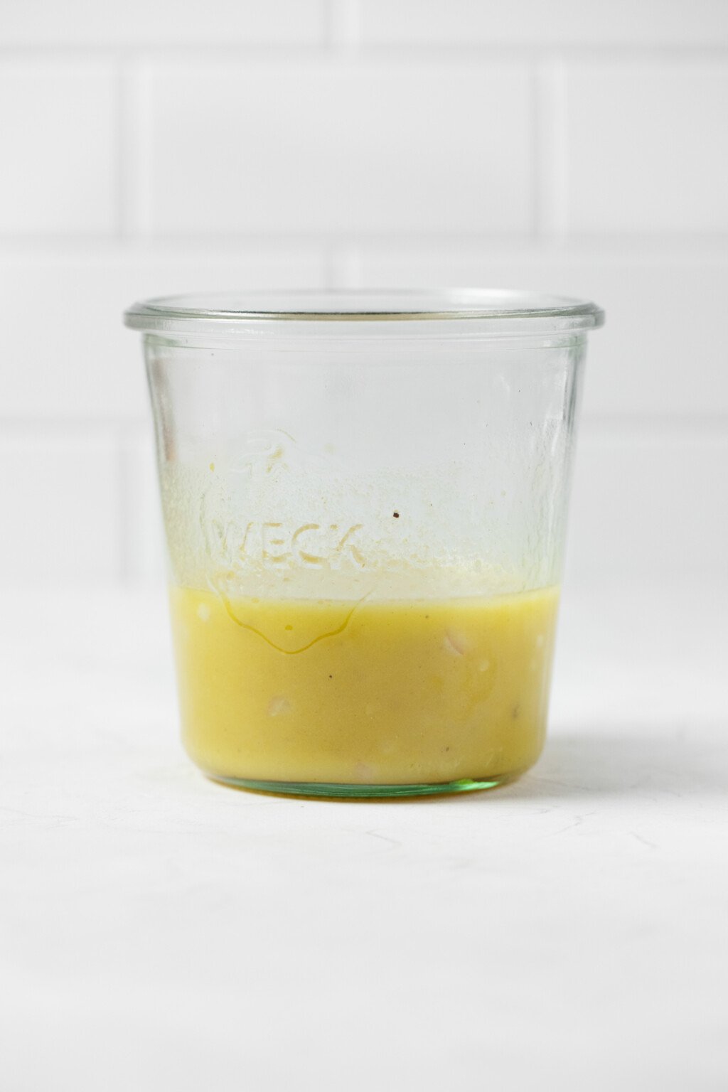A Weck mason jar, containing a creamy emulsified vinaigrette, rests on a white surface. There's a white tiled surface in the background.