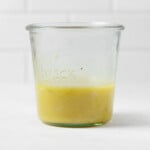 A Weck mason jar, containing a creamy emulsified vinaigrette, rests on a white surface. There's a white tiled surface in the background.