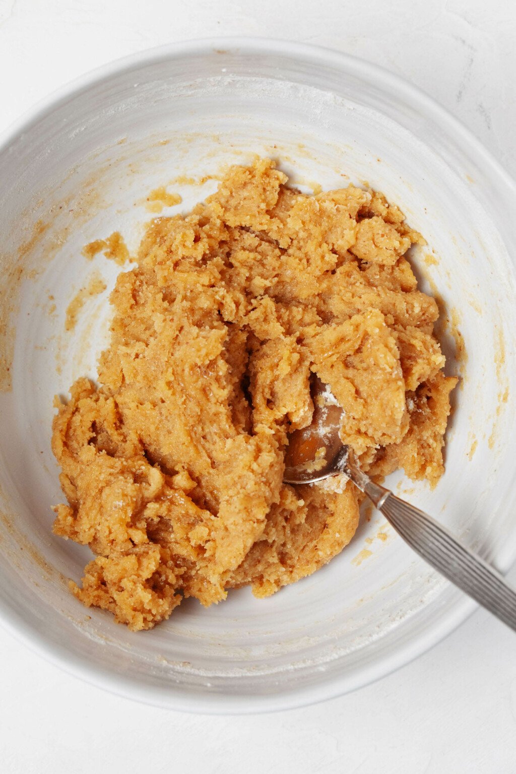 A white, round mixing bowl contains a light brown vegan cookie batter.