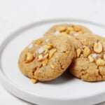 A round, rimmed white plate holds three vegan peanut cookies, each topped with chopped peanuts and flaky salt.