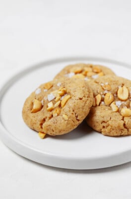 A round, rimmed white plate holds three vegan peanut cookies, each topped with chopped peanuts and flaky salt.