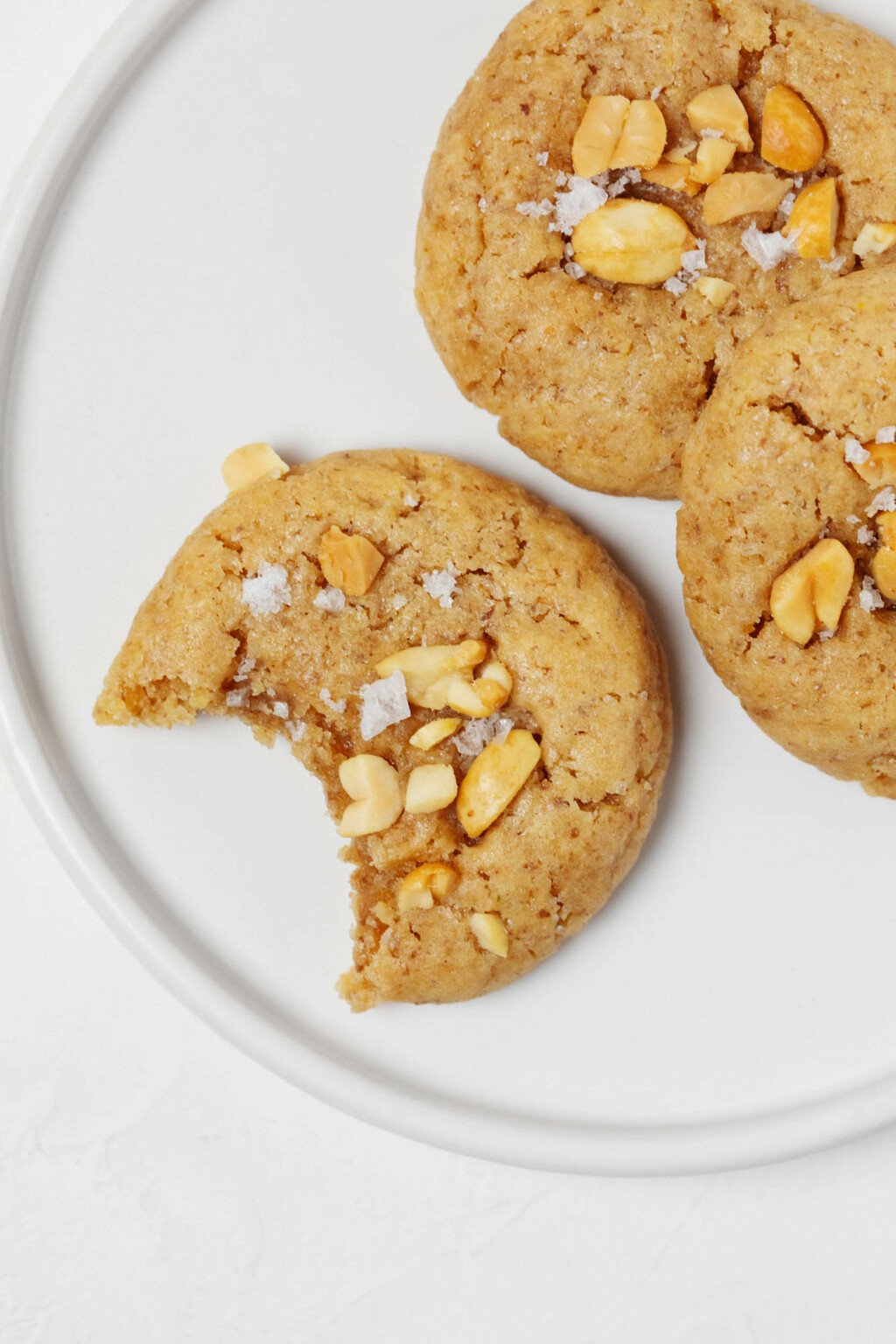 Three round, puffy vegan peanut cookies are positioned on a round, white plate. One of the cookies has a half-moon shaped bite taken out of it.
