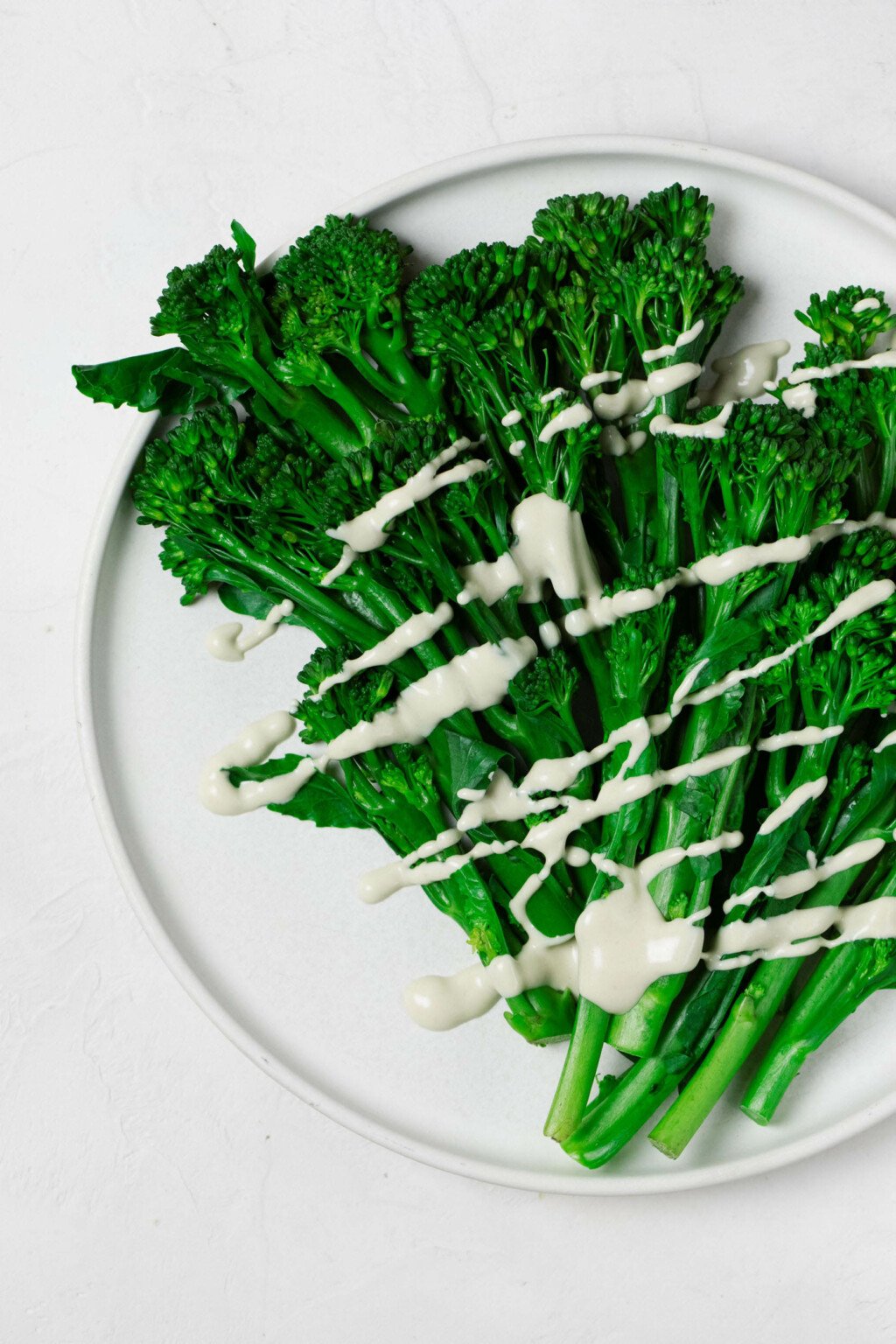 Ein Bild von oben: hellgrüner, gedämpfter Broccolini, der auf einem weißen, runden Teller ruht.  Der Broccolini wird mit einem cremig-weißen Tahini-Dressing beträufelt.