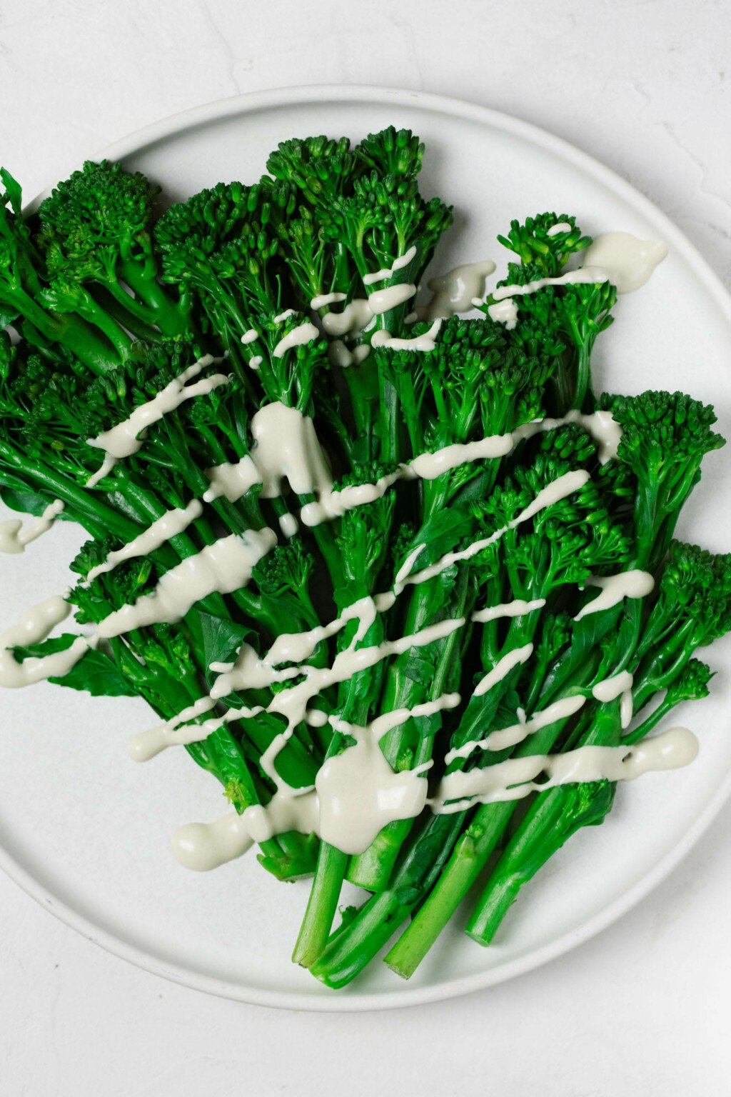 An overhead image of bright green, steamed broccolini resting on a white, round plate. The broccolini is drizzled with a creamy white tahini dressing.