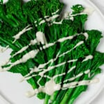 An overhead image of bright green, steamed broccolini resting on a white, round plate. The broccolini is drizzled with a creamy white tahini dressing.
