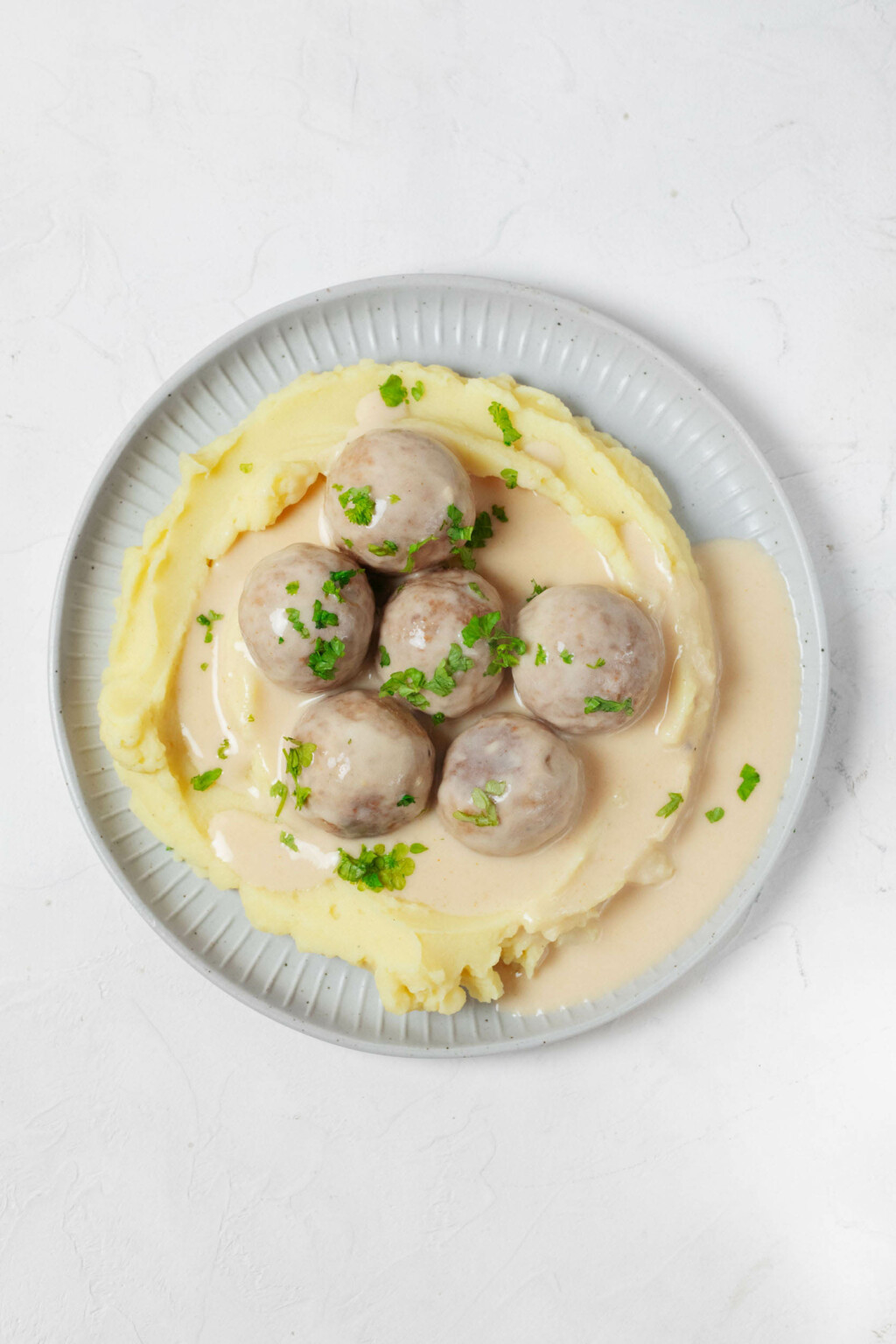 A round, gray white plate is topped with mashed potatoes, vegan Swedish meatballs, and chopped parsley.