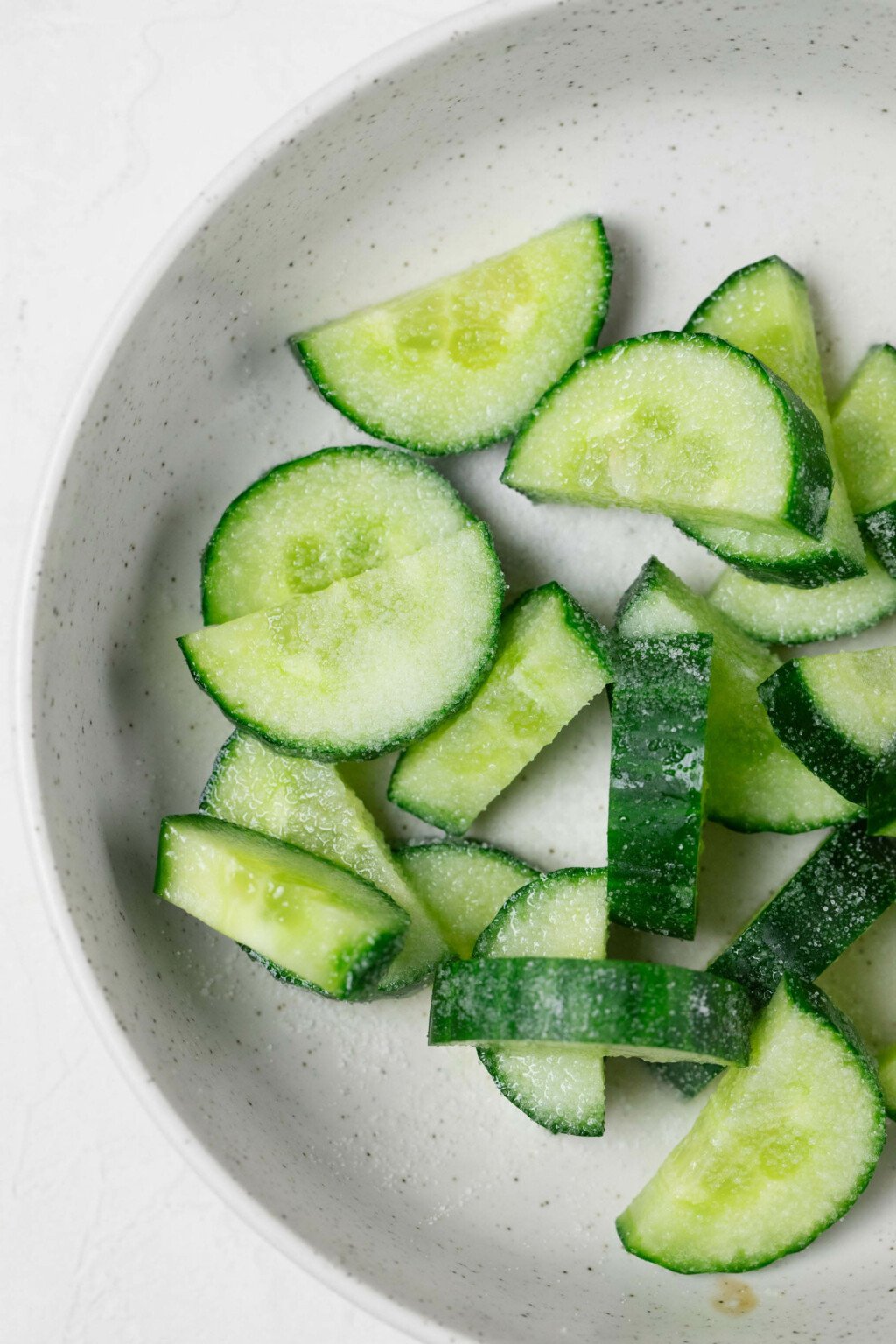An overhead image of a white bowl, filled with cut cucumber slices.