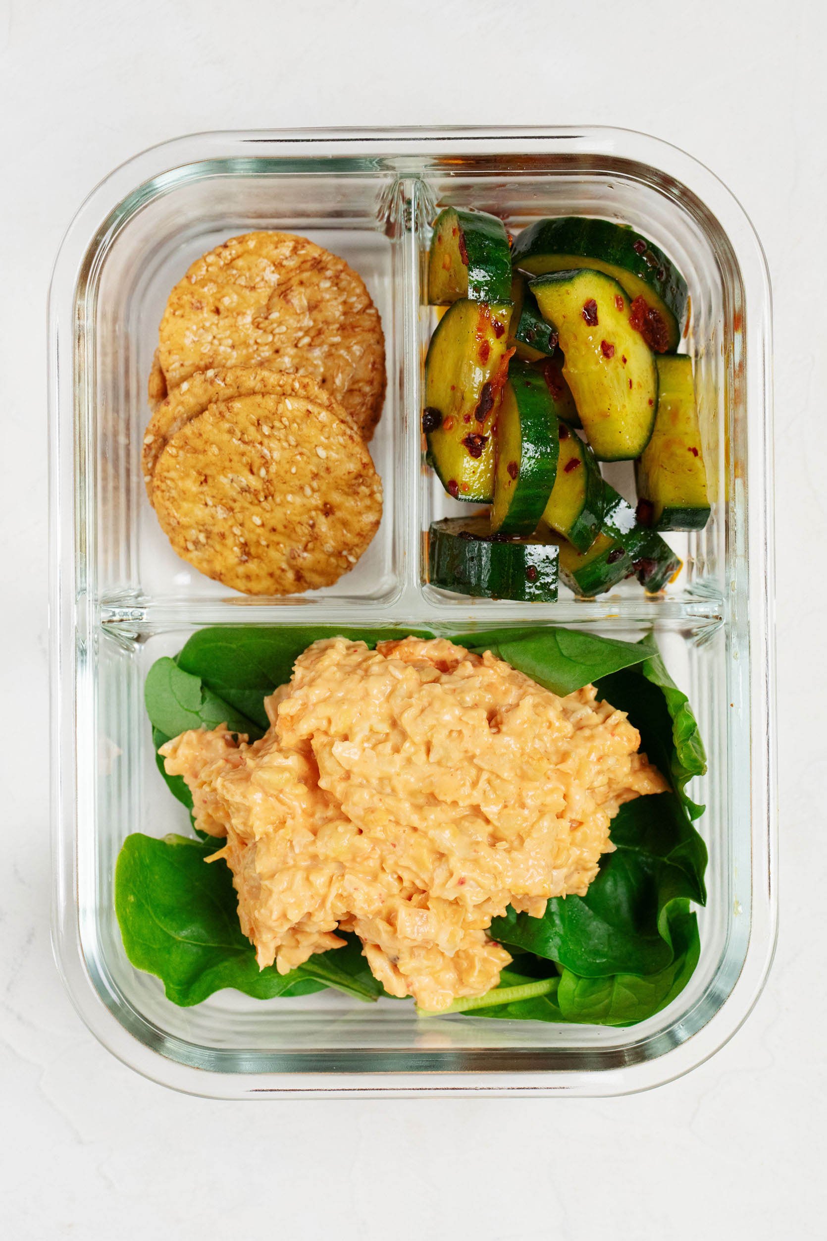 A small, Pyrex bento box is pictured on a white surface. It contains a smashed chickpea salad, marinated cucumbers, and rice crackers.