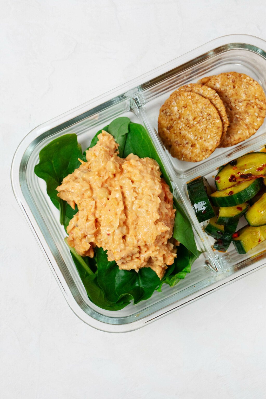 A small, Pyrex bento box is pictured on a white surface. It contains a smashed chickpea salad, marinated cucumbers, and rice crackers.