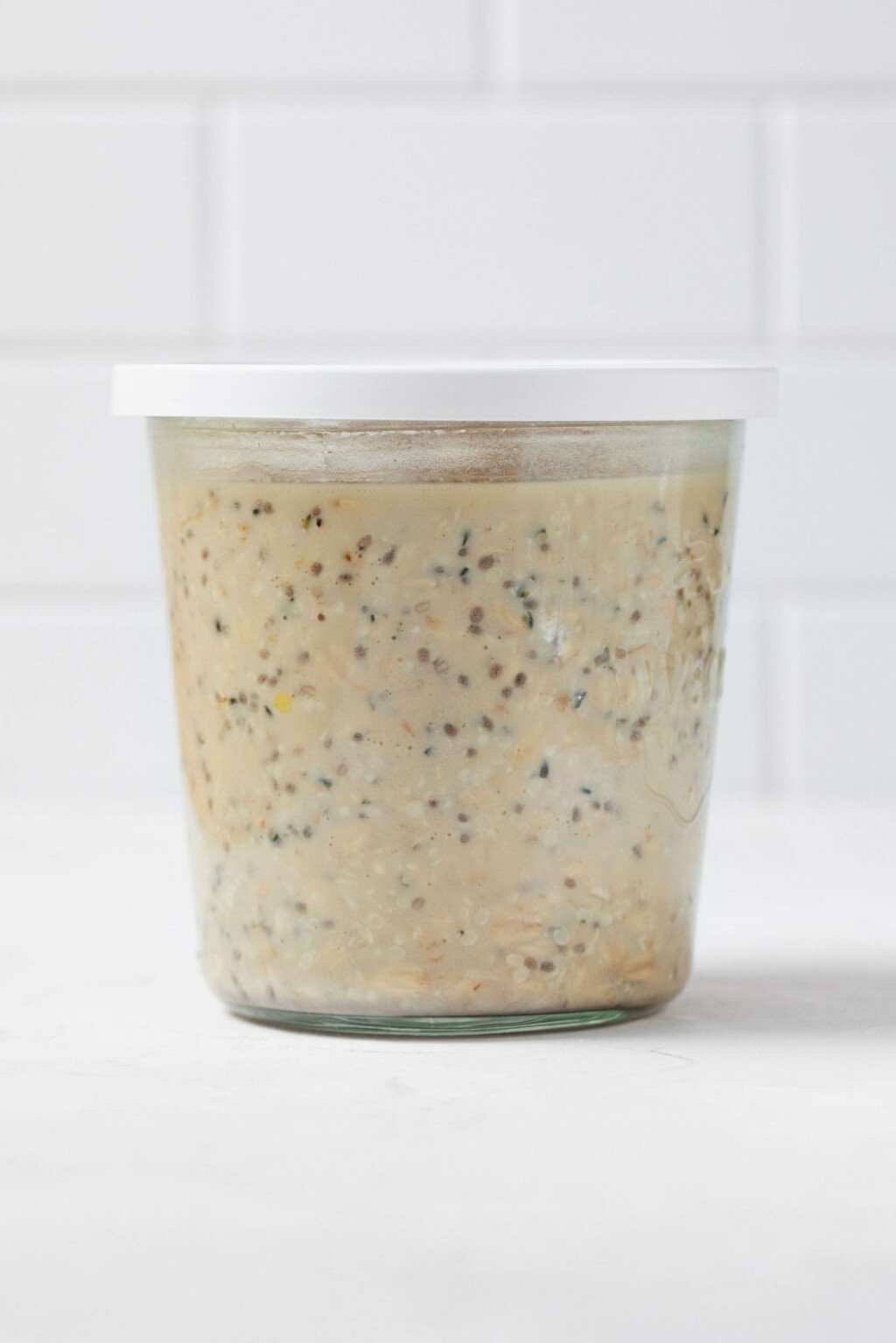 A glass mason jar is filled with vegan overnight oats. There's white brick visible in the background.