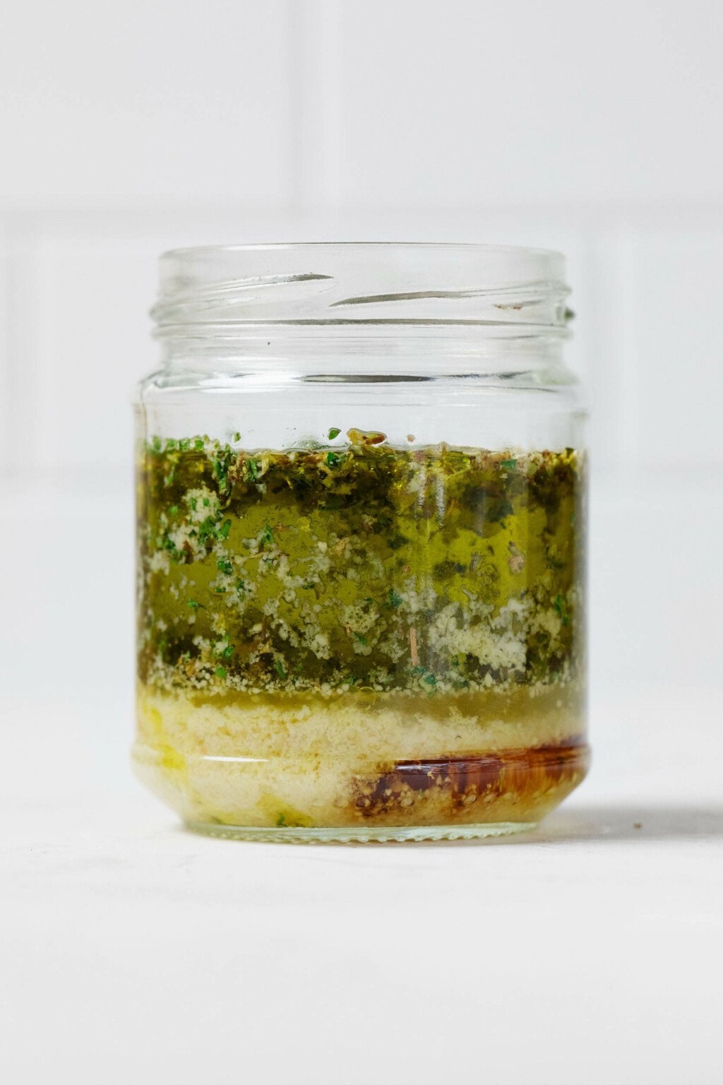 A glass gar is pictured angled, with a white surface in the background. The jar is filled with olive oil and other ingredients for a vinaigrette dressing.