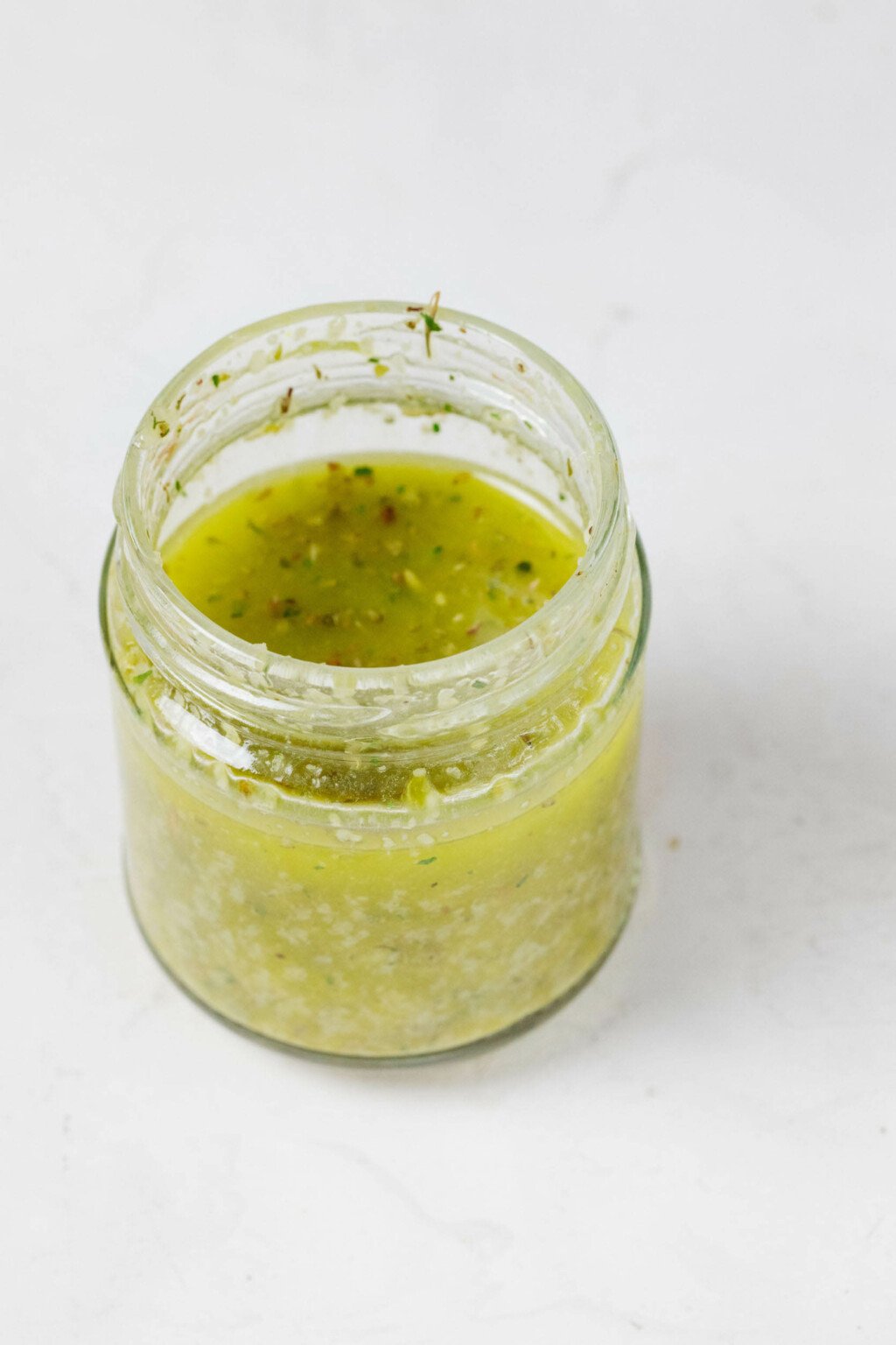 An angled image of a jar of salad dressing, which has been made with pale green olive oil. It rests on a white surface.