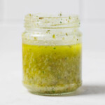An angled image of a jar of salad dressing, which has been made with pale green olive oil. It rests on a white surface.