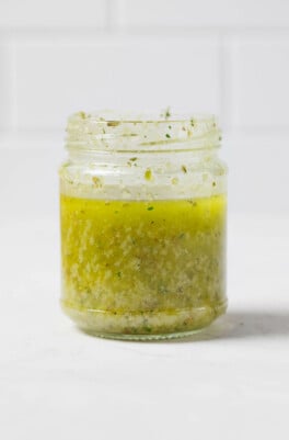 An angled image of a jar of salad dressing, which has been made with pale green olive oil. It rests on a white surface.