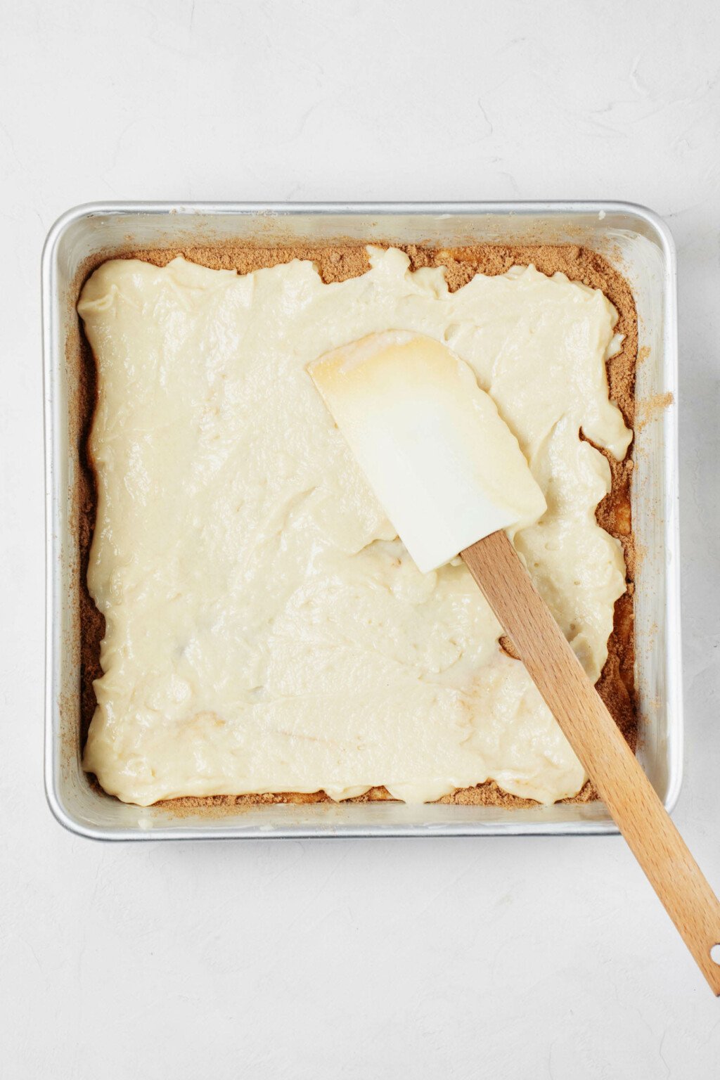 A spatula is being used to spread cake batter over a layer of cinnamon sugar in a square baking dish.