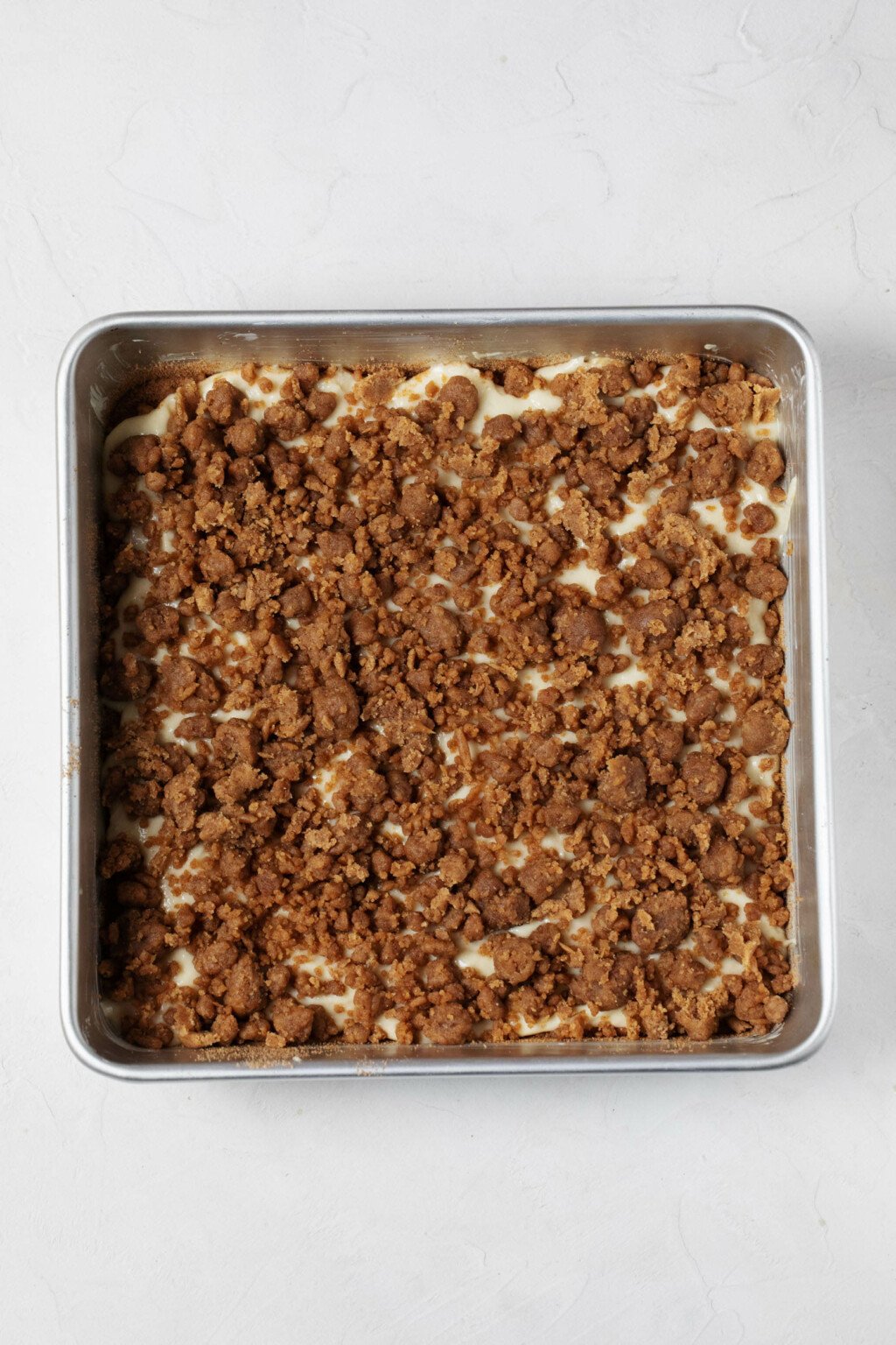 A square cake pan is pictured overhead. It contains a layer of cake batter topped with crumb-like streusel.