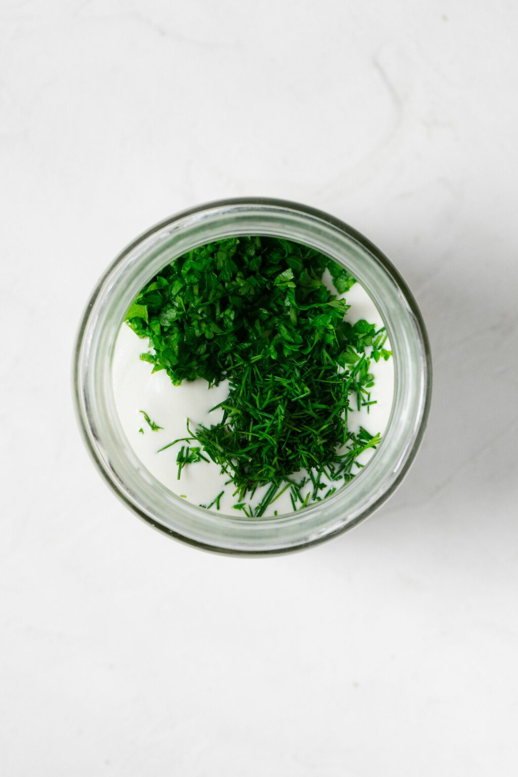 An overhead image of the round mouth of a glass jar. The jar is filled with a creamy dressing and fresh, chopped herbs.