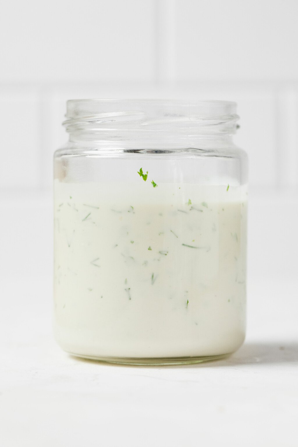 An angled image of a glass mason jar, resting on a white surface. It contains a creamy vegan ranch dressing with herbs.