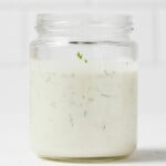 An angled image of a glass mason jar, resting on a white surface. It contains a creamy vegan ranch dressing with herbs.