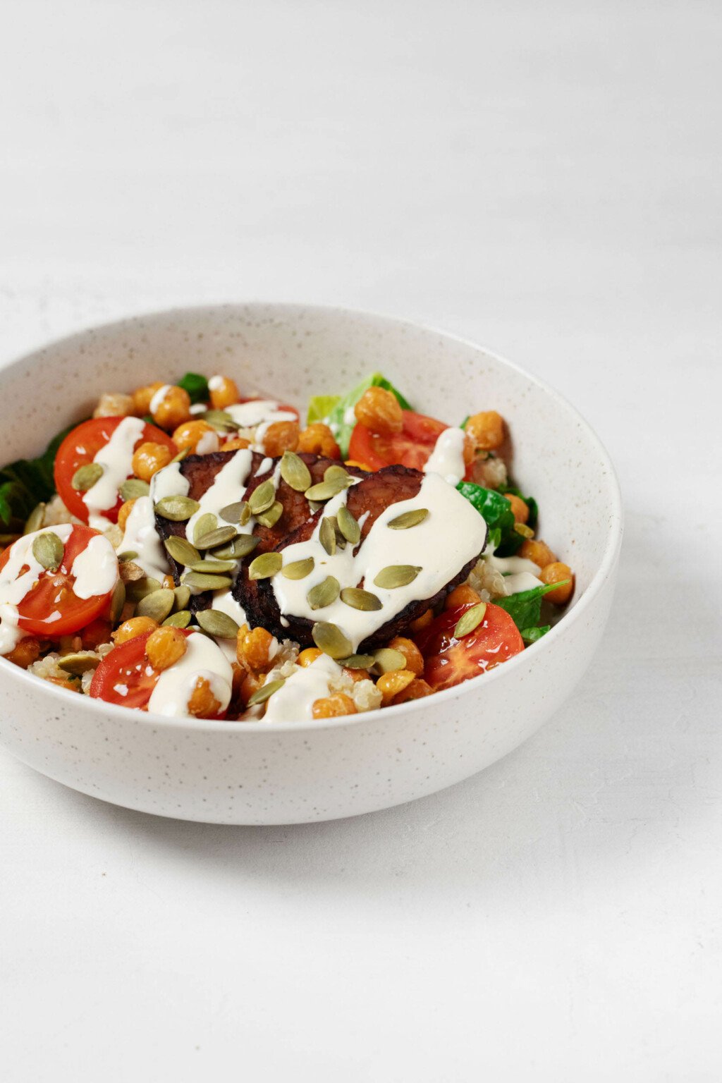 A large, white bowl is filled with vegetables, roasted chickpeas, quinoa, and a creamy ranch dressing. The bowl is topped with pepitas.