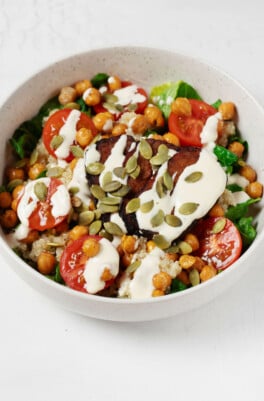 A large, white bowl is filled with vegetables, roasted chickpeas, quinoa, and a creamy ranch dressing. The bowl is topped with pepitas.