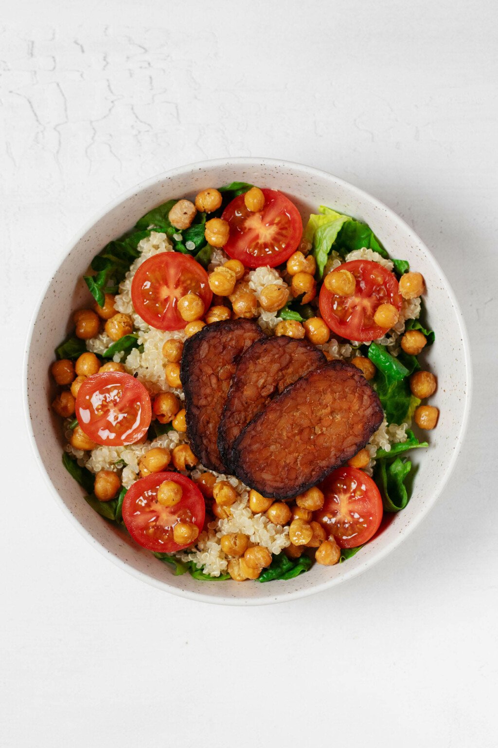 A white bowl holds greens, tomatoes, cooked quinoa, chickpeas, and some slices of tempeh bacon.