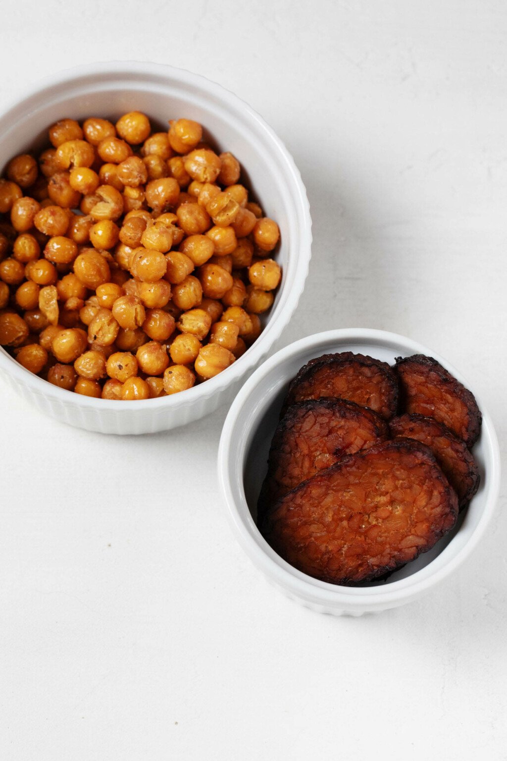 An overhead image of two small, white bowls. One contains crispy roasted chickpeas, while the other contains darkened, crispy baked tempeh bacon.
