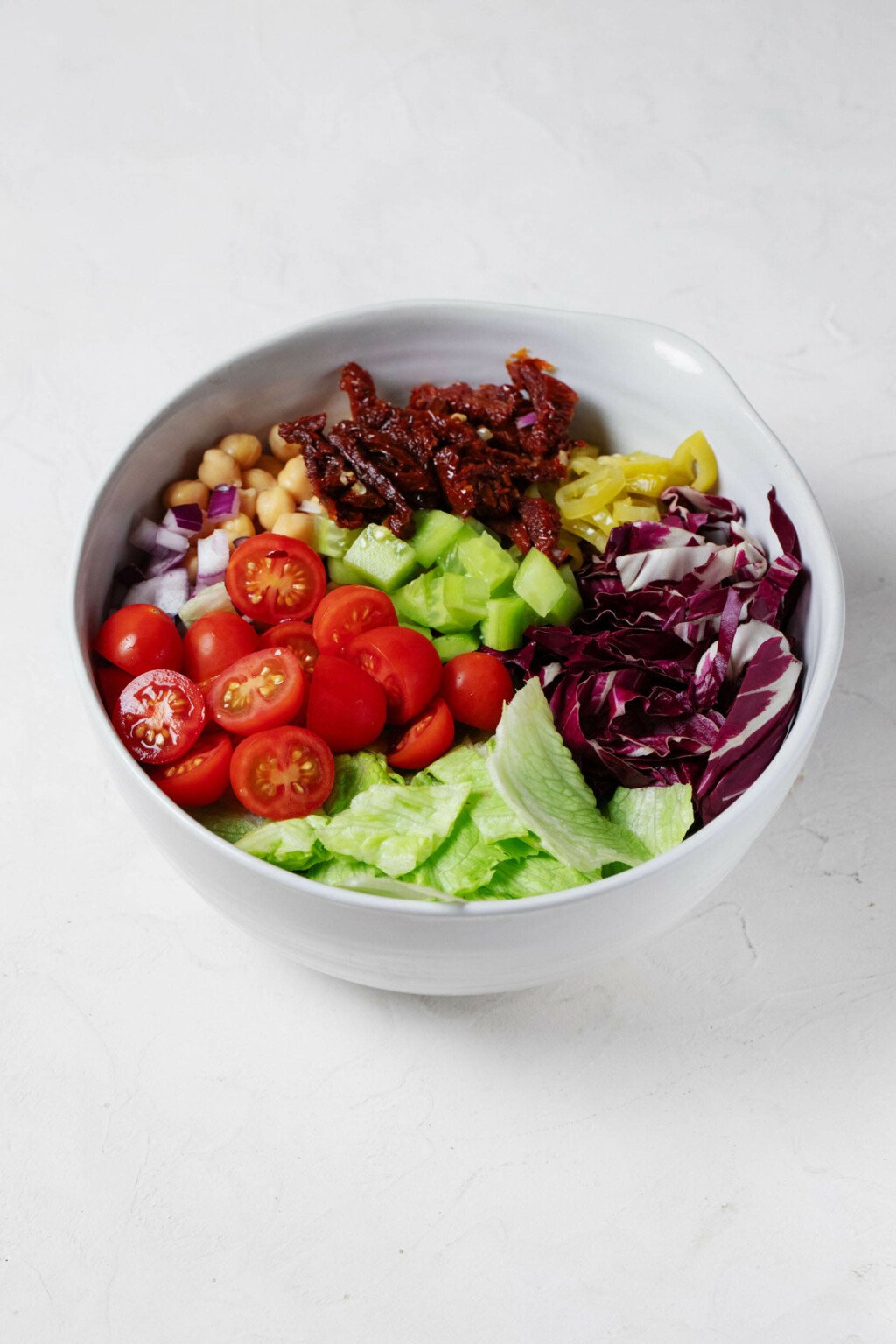 A white bowl is resting on a white surface. It's filled with a colorful mix of vegetables, including cucumber, radicchio, greens, and cherry tomatoes.