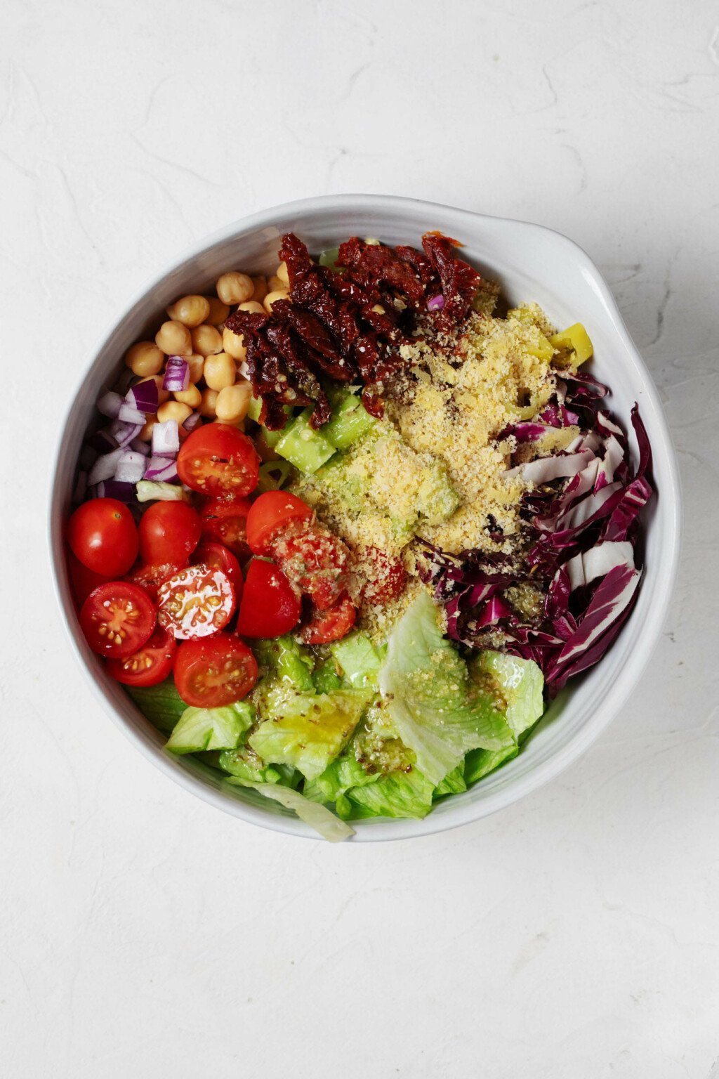 A white bowl is resting on a white surface. It's filled with a colorful mix of vegetables, including cucumber, radicchio, greens, and cherry tomatoes.