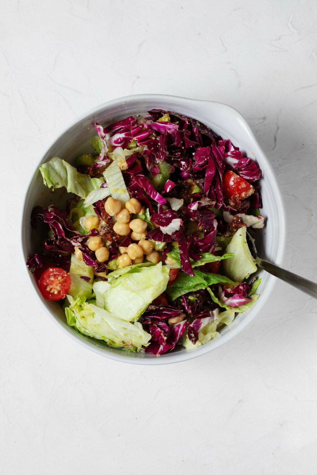 A white mixing bowl, which rests on a white surface, is holding the components for a colorful vegan Italian chopped salad with chickpeas.