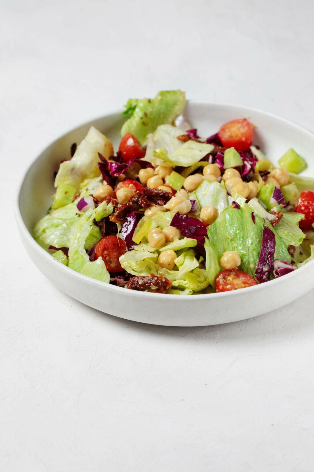 An angled image of a white bowl, which has been filled with a colorful Italian chopped salad mixture. It rests on a white surface.