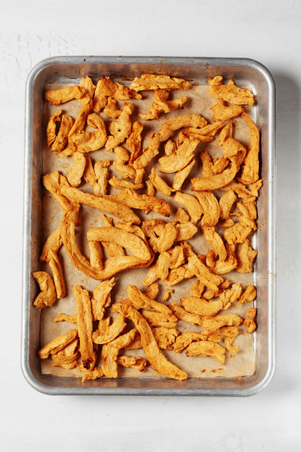 An overhead image of a metal baking sheet, which is topped with tan-colored vegan soy protein strips. 