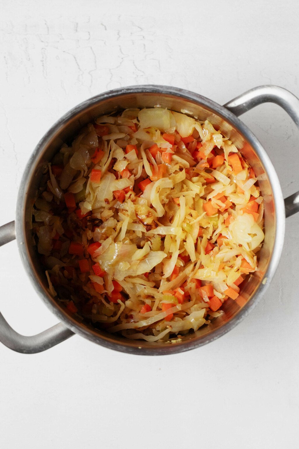 An overhead image of a silver pot with handles, which is being used to sauté cabbage, celery, and carrots. 