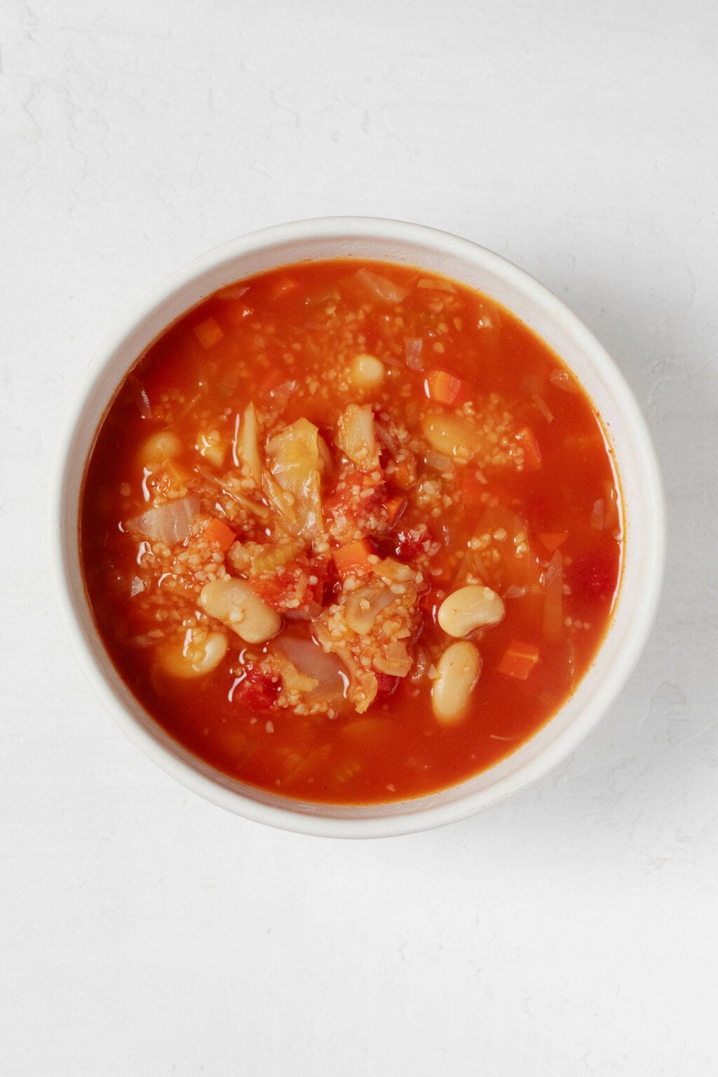 An overhead image of a white bowl of red-colored soup, which is filled with bulgur wheat and white beans.