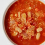 An overhead image of a white bowl of red-colored soup, which is filled with bulgur wheat and white beans.