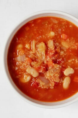 An overhead image of a white bowl of red-colored soup, which is filled with bulgur wheat and white beans.