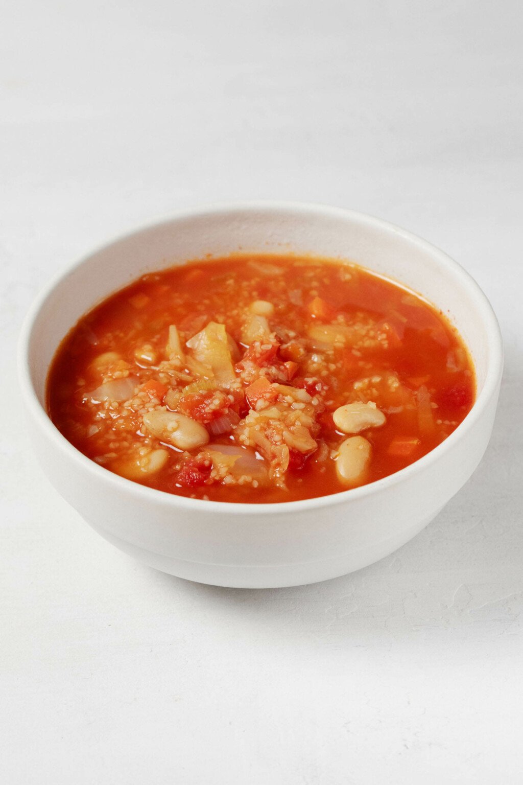 An image of a white bowl of red-colored soup, which is filled with bulgur wheat and white beans.