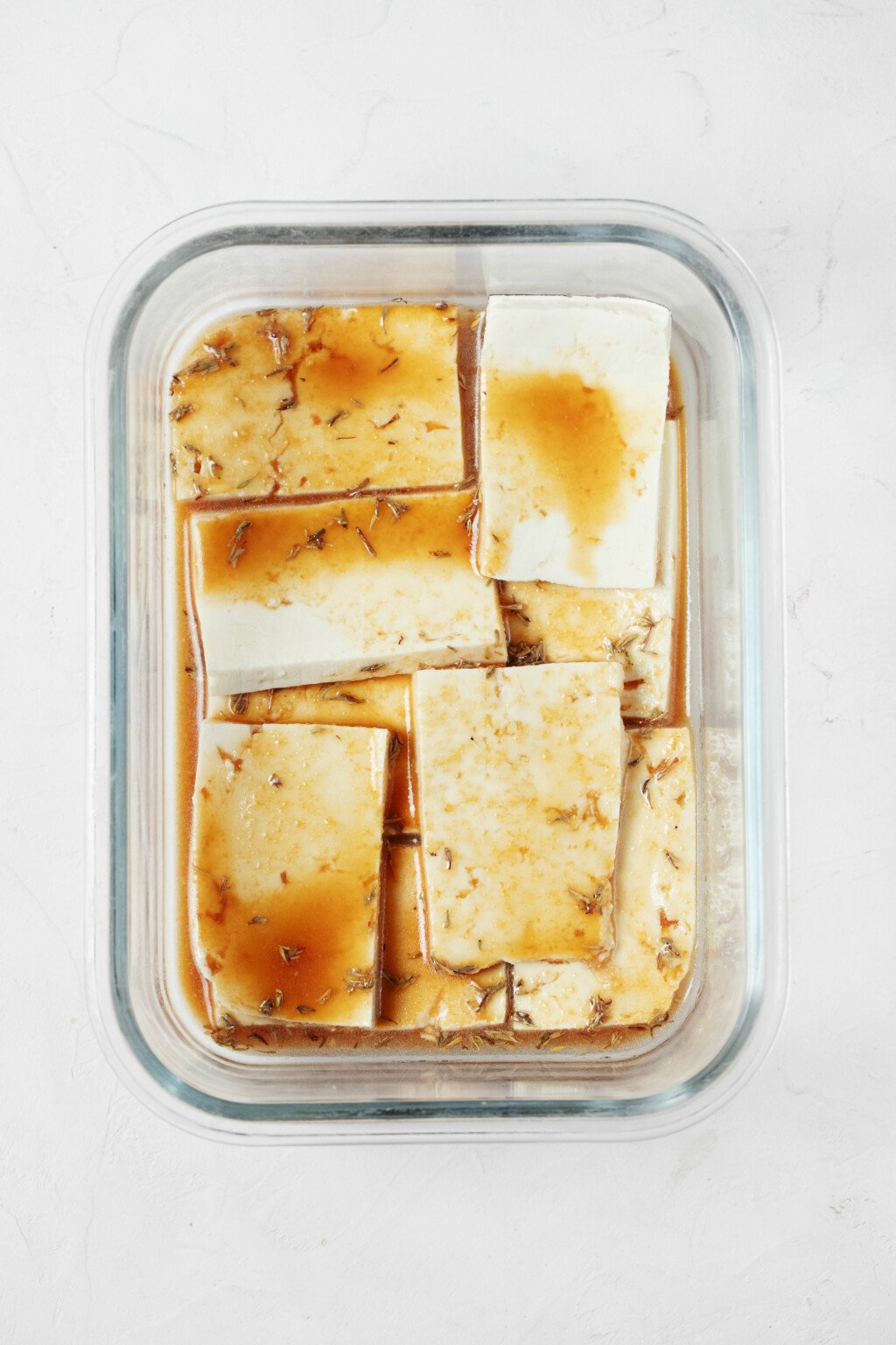 An overhead image of a glass storage container, in which slices of tofu have been piled and covered in marinade.